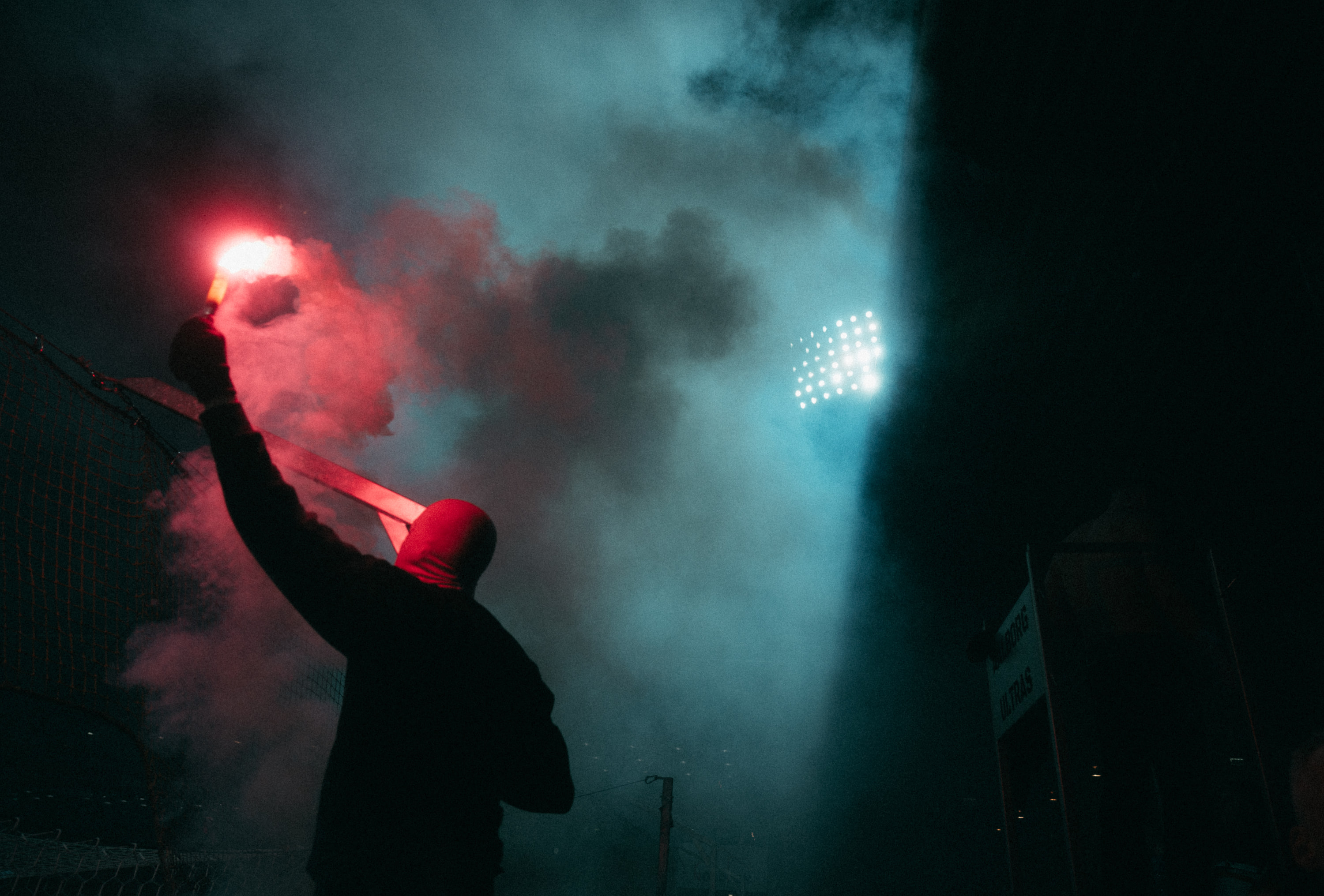 A silhouetted supporter raises a red flare beneath stadium floodlights