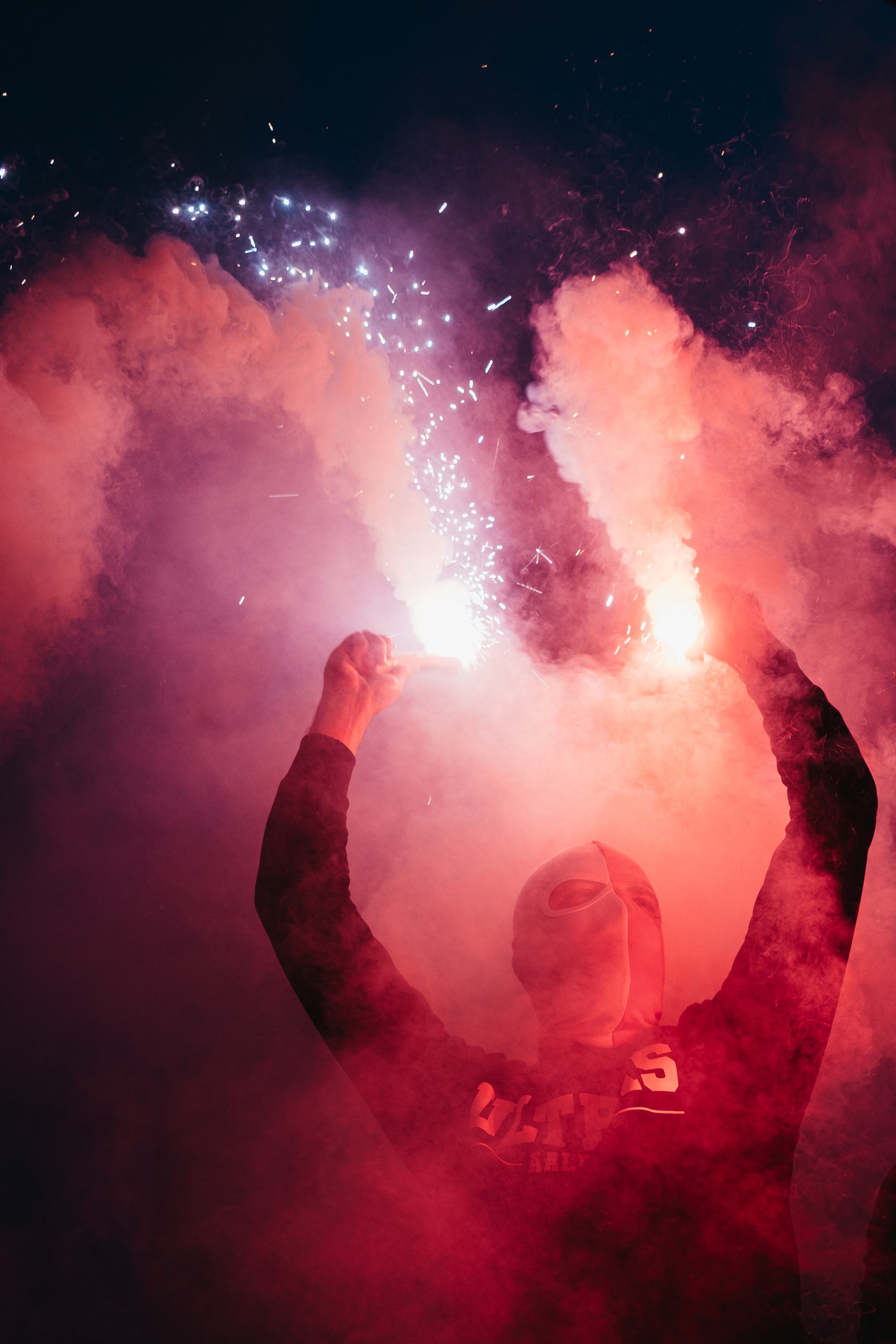 A masked supporter raises two flares through pink smoke