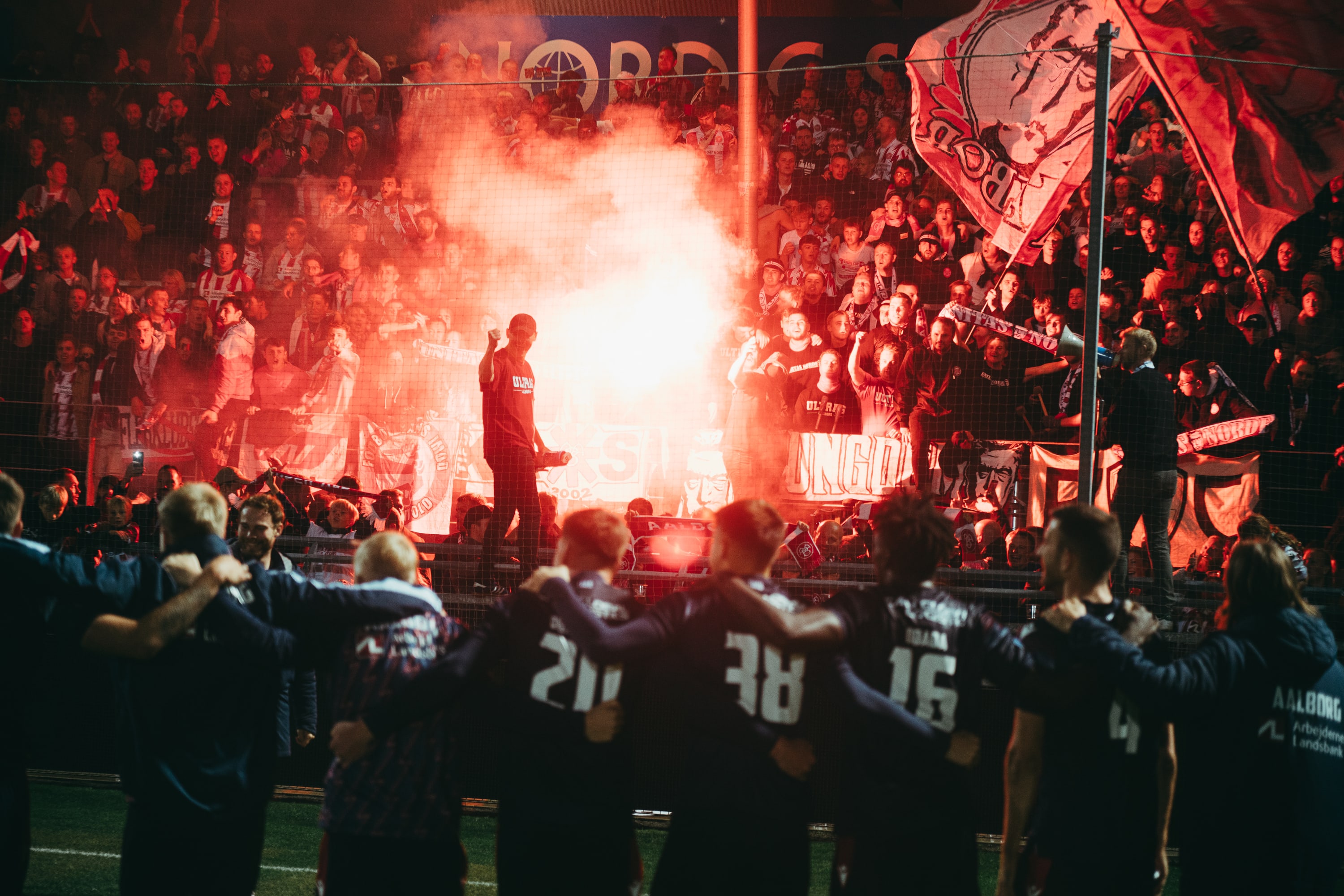 Football players face a stand of supporters illuminated by red flares