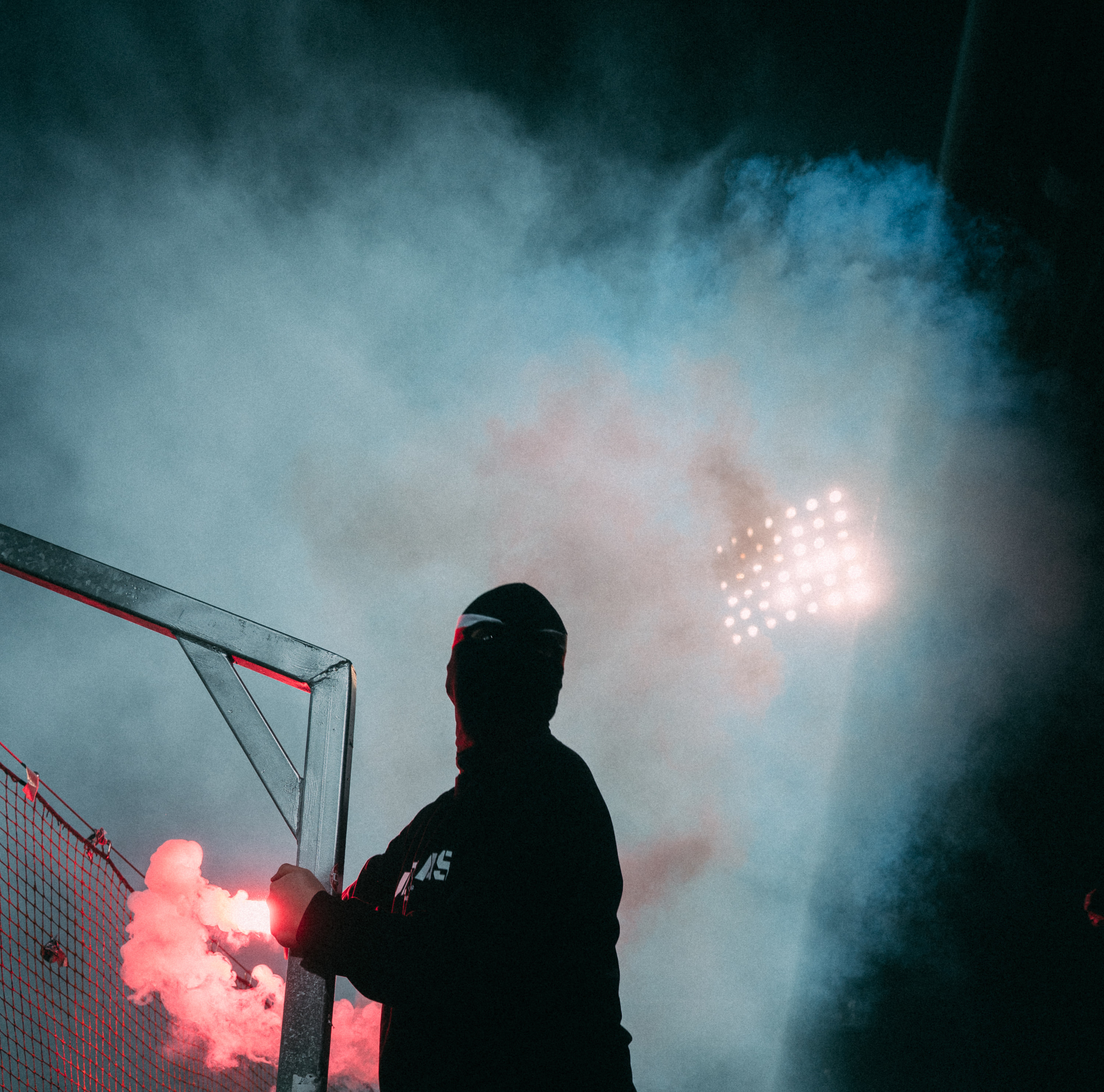 A masked supporter stands beside a stadium fence surrounded by flare smoke