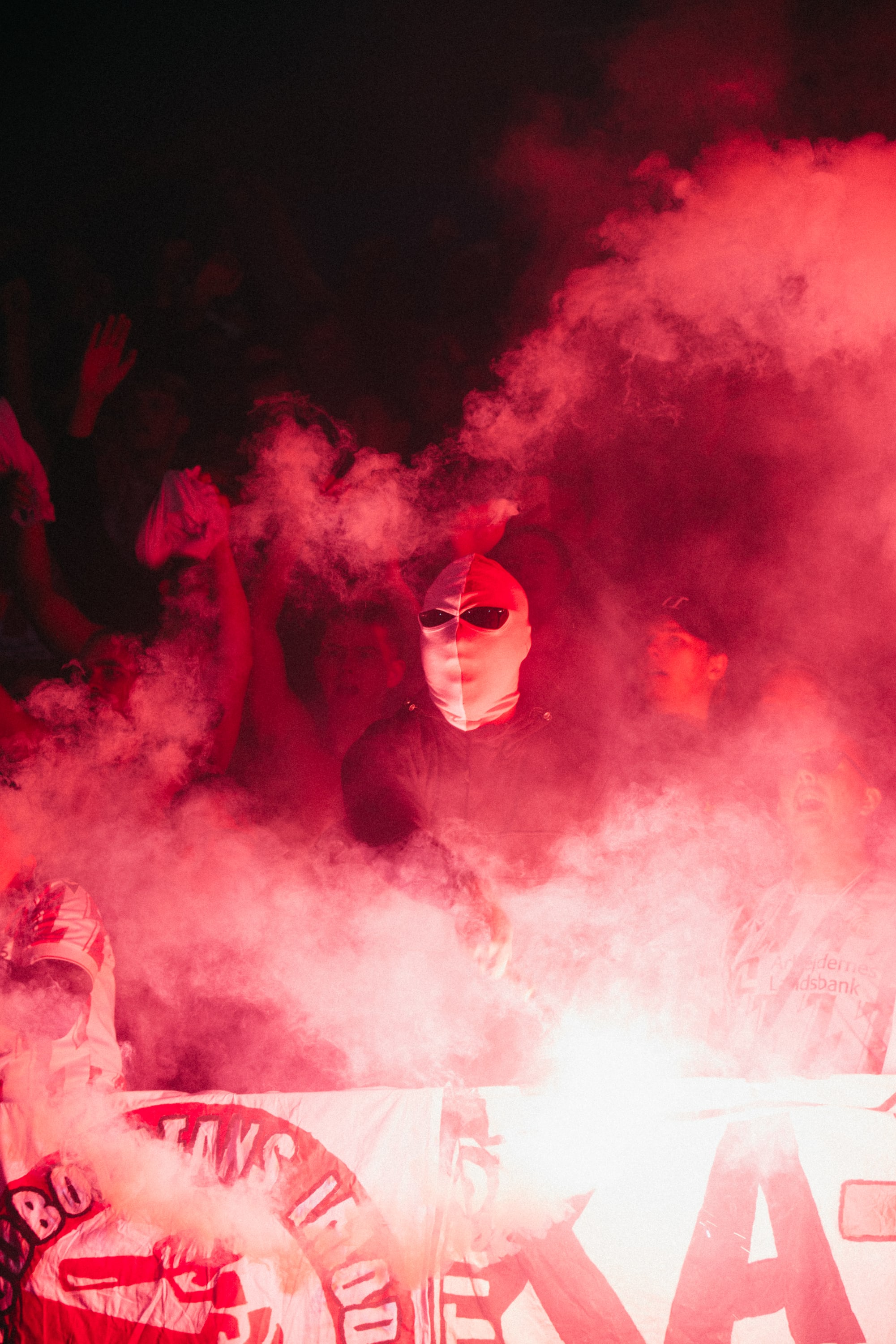 A masked supporter stands behind a banner in red flare smoke