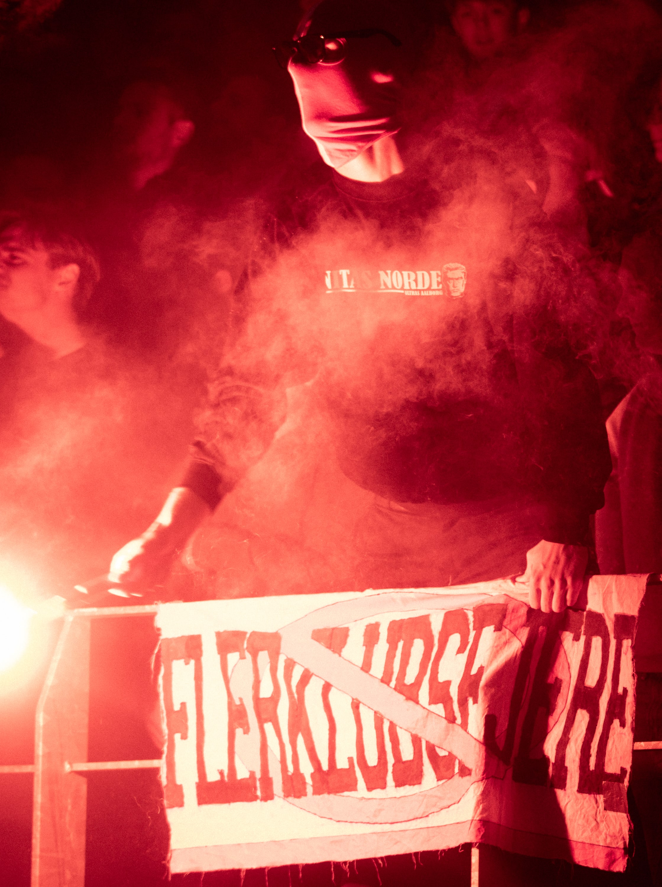 A masked supporter grips a banner in the glow of a red flare