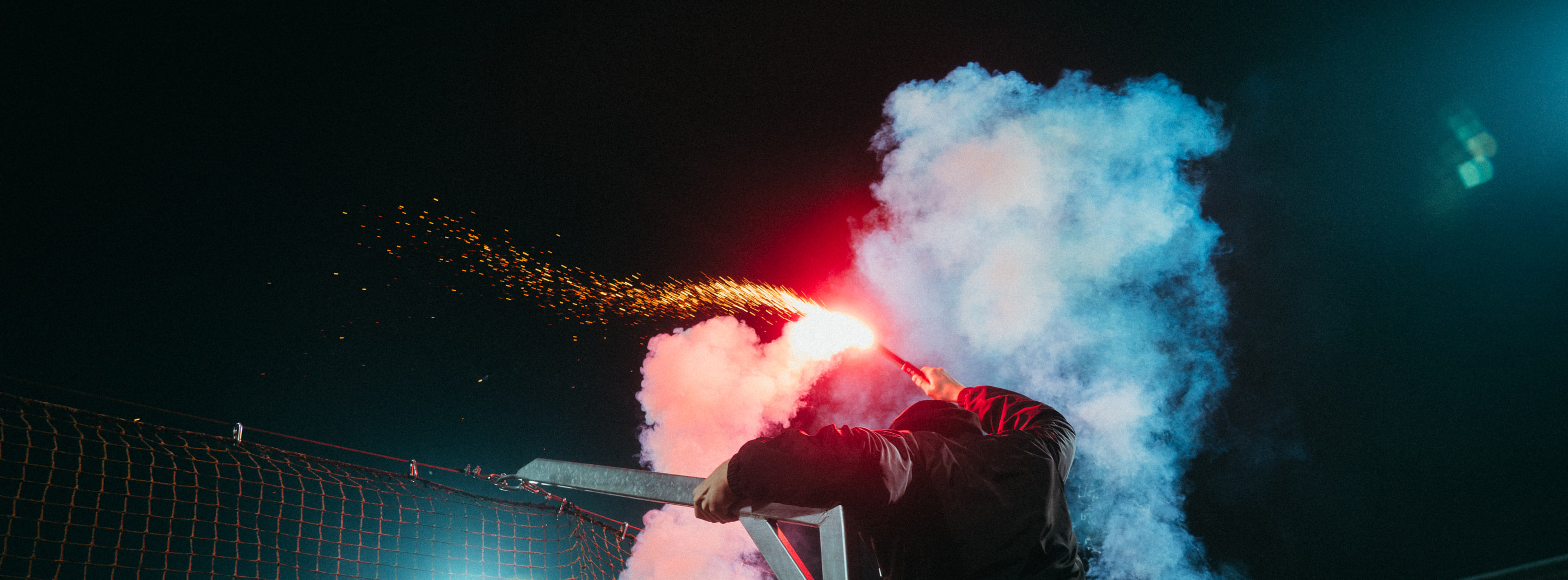 A masked supporter leans over a stadium fence with a burning red flare