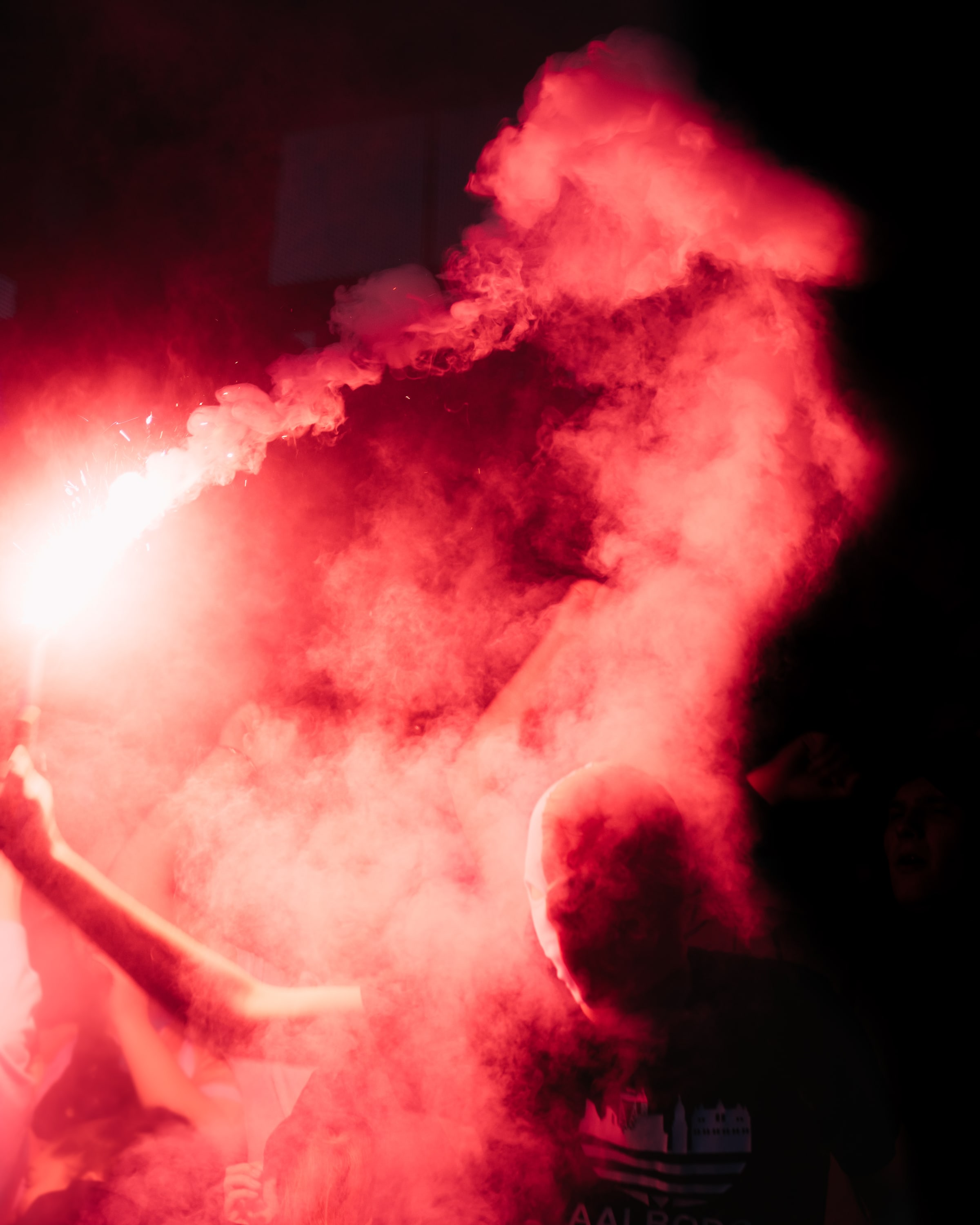 A masked supporter holds a burning flare amid dense red smoke